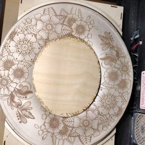 Puede incluir: Una pieza de madera redonda con un diseño floral grabado en su superficie. El diseño presenta flores y hojas detalladas. La pieza está colocada dentro de una máquina de grabado láser, mostrando el proceso de creación.