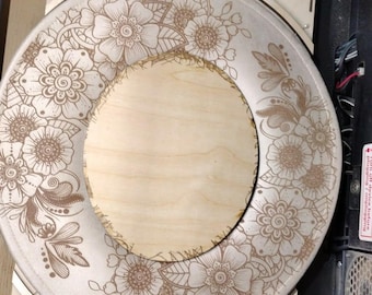 May include: A round wooden piece with a floral design engraved on its surface. The design features detailed flowers and leaves. The piece is placed inside a laser engraving machine, showing the process of creation.