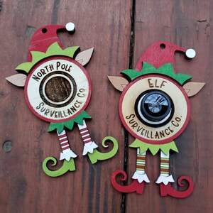 May include: Two wooden elf ornaments with red hats and green elf ears. The ornaments have a round wooden circle with the text "North Pole Surveillance Co." and "Elf Surveillance Co."  The ornaments are hanging on a wooden surface.