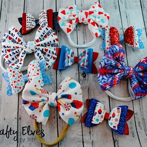May include: A collection of red, white, and blue hair bows with various patriotic designs, including glitter, stars, and stripes. The bows are displayed on a wooden surface with the text "Krafty Elves Est 2019".