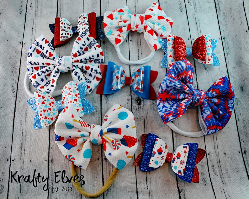 May include: A collection of red, white, and blue hair bows with various patriotic designs, including glitter, stars, and stripes. The bows are displayed on a wooden surface with the text "Krafty Elves Est 2019".