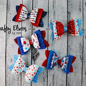 May include: Five red, white, and blue glitter hair bows with various patterns. The bows are arranged on a white wood background. The text "Krafty Elves Est. 2019" is visible in the image.