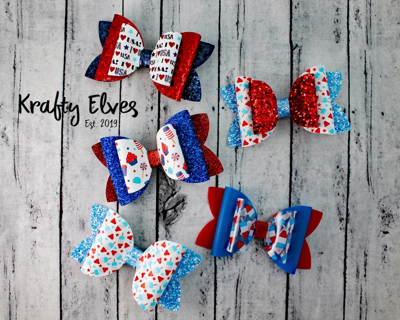 May include: Five red, white, and blue glitter hair bows with various patterns. The bows are arranged on a white wood background. The text "Krafty Elves Est. 2019" is visible in the image.