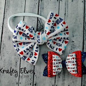 May include: Two hair bows with a red, white, and blue "I Love USA" print. The larger bow is white with blue stars and red hearts. The smaller bow is red with blue glitter and white stars. Both bows are attached to a white elastic headband.