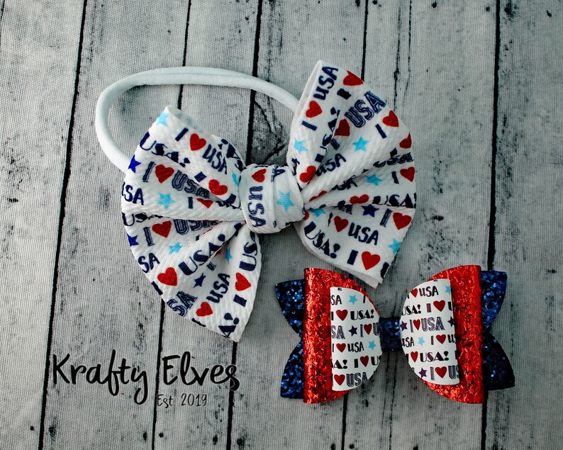 May include: Two hair bows with a red, white, and blue "I Love USA" print. The larger bow is white with blue stars and red hearts. The smaller bow is red with blue glitter and white stars. Both bows are attached to a white elastic headband.