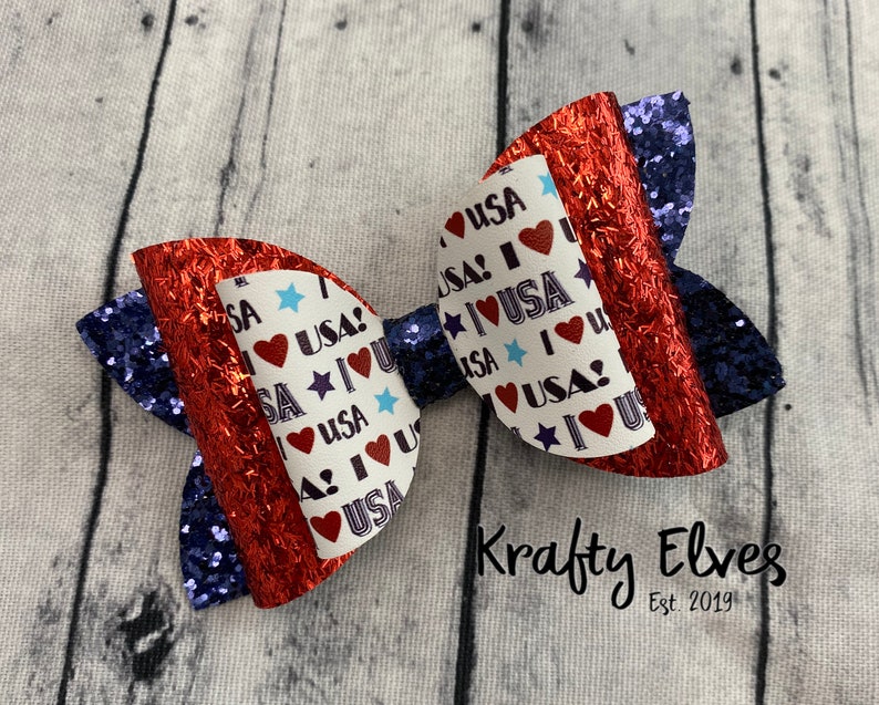 May include: Red, white, and blue glitter bow with a "I Love USA" print. The bow is made of fabric and has a glittery finish.