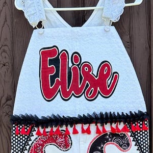 May include: White apron with the name "Elise" in red script with a black outline. The apron has eyelet lace detailing, black trim, and red tassels. The bottom features a paisley pattern.