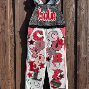 May include: White overalls featuring black and white floral bib with the name Caroline in red script. The legs are decorated with red, white, and black fabric appliqués spelling Coppell Cowboys, accented with stars and a small red cowboy hat.