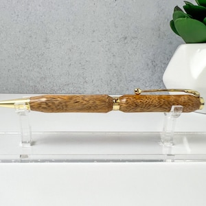 May include: A wooden pen with a gold-colored clip and accents. The pen is resting on a clear acrylic stand.