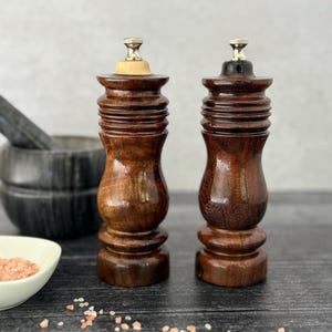 May include: Two wooden salt and pepper grinders with a rich, dark brown finish. The grinders have a classic shape with a decorative top and are set on a dark wooden surface. A small bowl of pink salt and a mortar and pestle are in the background.