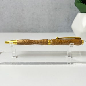 May include: A wooden pen with a gold-toned finish and a brown wood grain pattern. The pen is displayed on a clear acrylic stand.