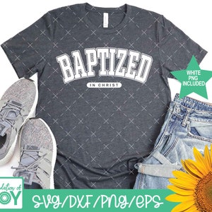 Puede incluir: Una camiseta gris oscuro con el texto "BAPTIZED IN CHRIST" en letras blancas. La camiseta est&aacute; sobre un fondo blanco con un par de zapatillas grises, unos vaqueros azules y un girasol.