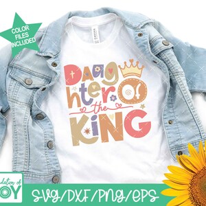 May include: A white t-shirt with the text "Daughter of the King" in colorful letters with a crown and hearts. The shirt is on a denim jacket and a sunflower.
