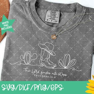 K&ouml;nnte beinhalten: Ein graues Comfort Colors T-Shirt mit einer wei&szlig;en Strichzeichnung eines Cowboyhuts, Stiefeln und W&uuml;stenpflanzen. Der Text "The Lord guides our steps" steht darunter. Eine goldene Halskette und ein brauner Hut sind ebenfalls zu sehen.