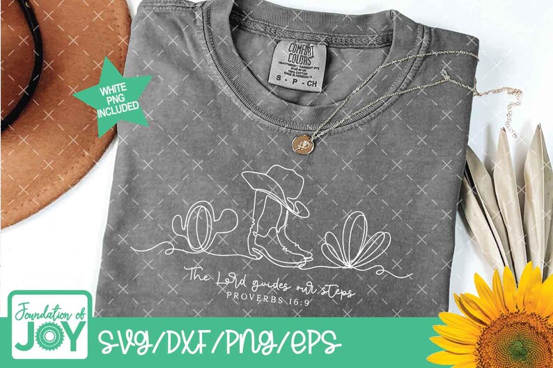 K&ouml;nnte beinhalten: Ein graues Comfort Colors T-Shirt mit einer wei&szlig;en Strichzeichnung eines Cowboyhuts, Stiefeln und W&uuml;stenpflanzen. Der Text "The Lord guides our steps" steht darunter. Eine goldene Halskette und ein brauner Hut sind ebenfalls zu sehen.