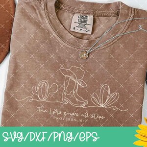 K&ouml;nnte beinhalten: Braunes T-Shirt mit einer wei&szlig;en Strichzeichnung eines Cowboyhuts, Stiefeln und Kakteen. Der Text "The Lord guides our steps PROVERBS 16:9" befindet sich darunter. Eine silberne Halskette und eine Sonnenblume sind ebenfalls zu sehen.