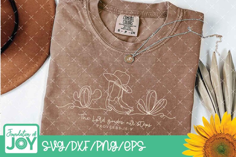 K&ouml;nnte beinhalten: Braunes T-Shirt mit einer wei&szlig;en Strichzeichnung eines Cowboyhuts, Stiefeln und Kakteen. Der Text "The Lord guides our steps PROVERBS 16:9" befindet sich darunter. Eine silberne Halskette und eine Sonnenblume sind ebenfalls zu sehen.