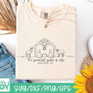 May include: Cream-colored t-shirt with a black line drawing of a nativity scene, including a donkey, figures, and a sheep. The text "The greatest gift of all" and "Matthew 1:21" are below the drawing. A gold necklace and a Comfort Colors tag are visible.
