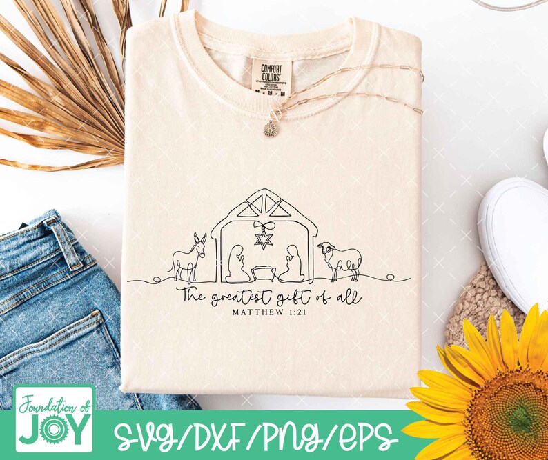 May include: Cream-colored t-shirt with a black line drawing of a nativity scene, including a donkey, figures, and a sheep. The text "The greatest gift of all" and "Matthew 1:21" are below the drawing. A gold necklace and a Comfort Colors tag are visible.