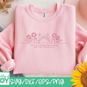 May include: A light pink sweatshirt with a crew neck and long sleeves. The design features a line drawing of sunflowers, butterflies, and a sun, with the text "When you can't find the sunshine, be the sunshine."