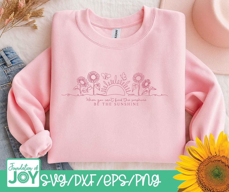 May include: A light pink sweatshirt with a crew neck and long sleeves. The design features a line drawing of sunflowers, butterflies, and a sun, with the text "When you can't find the sunshine, be the sunshine."