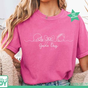 May include: A bright pink short-sleeve t-shirt featuring a white continuous line drawing of a football helmet, a bow, and a football. Below the graphic, the text Game Day is written in a white cursive font. A green star icon says WHITE PNG INCLUDED.