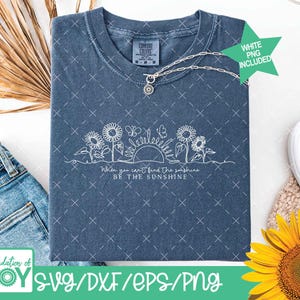 May include: A blue t-shirt with a white floral design featuring sunflowers, butterflies, and the sun. The text reads "When you can't find the sunshine, be the sunshine." A silver necklace with a sunflower pendant is also visible.