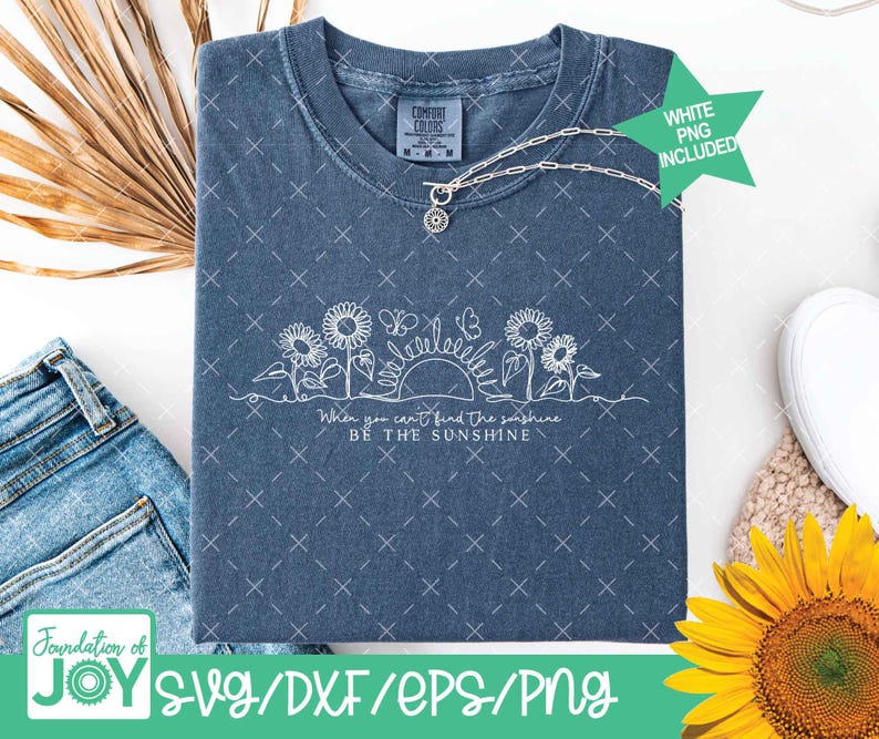 May include: A blue t-shirt with a white floral design featuring sunflowers, butterflies, and the sun. The text reads "When you can't find the sunshine, be the sunshine." A silver necklace with a sunflower pendant is also visible.