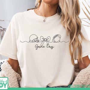 May include: A cream-colored short-sleeve t-shirt featuring a black continuous line drawing of a football helmet, a bow, and a football. Below the graphic, the text Game Day is written in a black cursive script. The shirt has a crew neckline.