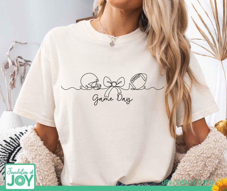 May include: A cream-colored short-sleeve t-shirt featuring a black continuous line drawing of a football helmet, a bow, and a football. Below the graphic, the text Game Day is written in a black cursive script. The shirt has a crew neckline.