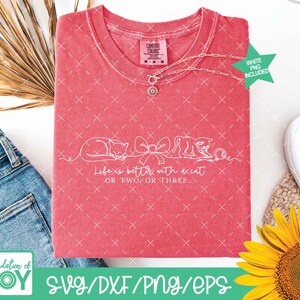 May include: A coral-colored t-shirt with a white graphic of two cats and a bow, with the text "Life is better with a cat or two or three." A silver necklace with a pendant is draped across the shirt. The shirt is next to a pair of blue jeans and a sunflower.