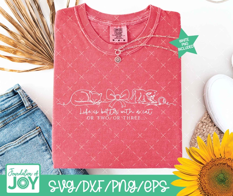 May include: A coral-colored t-shirt with a white graphic of two cats and a bow, with the text "Life is better with a cat or two or three." A silver necklace with a pendant is draped across the shirt. The shirt is next to a pair of blue jeans and a sunflower.