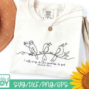 May include: Cream-colored t-shirt featuring a black line drawing of three singing birds perched on a branch. The text "I will sing of the goodness of god, Psalm 89:1" is printed below the birds. A gold necklace is draped on the shirt.