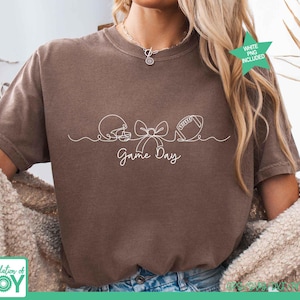 May include: A brown short-sleeve t-shirt featuring a white continuous line drawing of a football helmet, a bow, and a football. Below the graphic, the text Game Day is printed in a white cursive script. The shirt has a crew neckline.