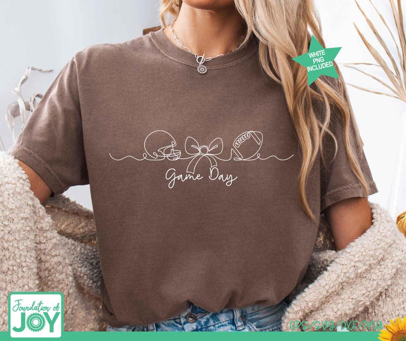 May include: A brown short-sleeve t-shirt featuring a white continuous line drawing of a football helmet, a bow, and a football. Below the graphic, the text Game Day is printed in a white cursive script. The shirt has a crew neckline.