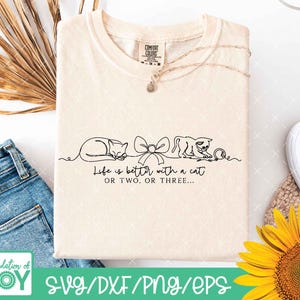 May include: Cream-colored t-shirt with a black line drawing of two cats, a bow, and a ball of yarn. The text "Life is better with a cat OR TWO. OR THREE..." is printed below the drawing. A silver necklace is draped across the shirt.