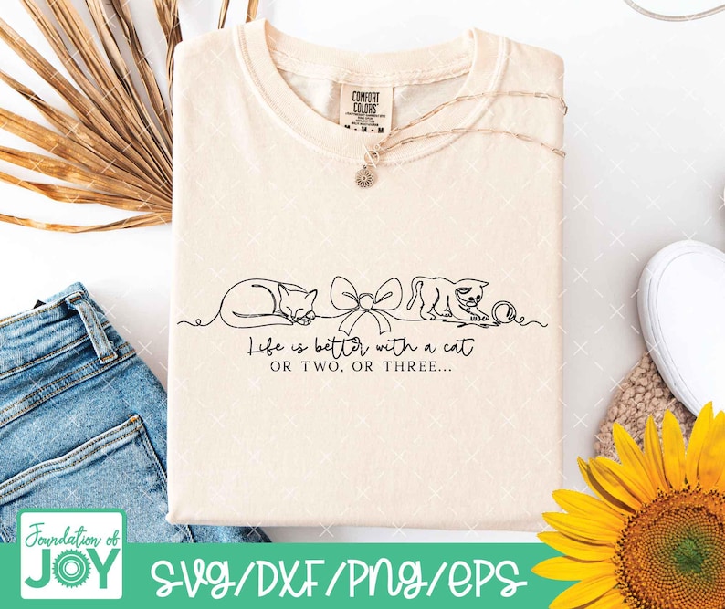 May include: Cream-colored t-shirt with a black line drawing of two cats, a bow, and a ball of yarn. The text "Life is better with a cat OR TWO. OR THREE..." is printed below the drawing. A silver necklace is draped across the shirt.
