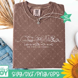 May include: A brown Comfort Colors t-shirt with a white line drawing of two cats and a bow. The text "Life is better with a cat or two or three" is below the drawing. A silver necklace with a charm is on the shirt.