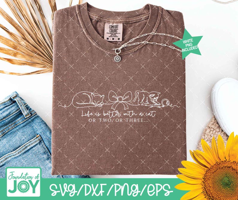 May include: A brown Comfort Colors t-shirt with a white line drawing of two cats and a bow. The text "Life is better with a cat or two or three" is below the drawing. A silver necklace with a charm is on the shirt.