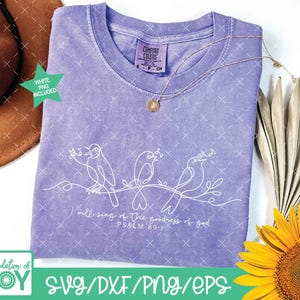 May include: A light purple t-shirt with a white line drawing of three birds on a branch. The text "I will sing of the goodness of God, Psalm 89:1" is below the birds. A gold necklace and a sunflower are also in the image.