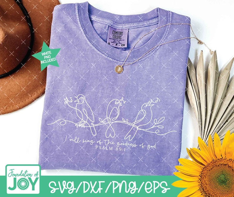 May include: A light purple t-shirt with a white line drawing of three birds on a branch. The text "I will sing of the goodness of God, Psalm 89:1" is below the birds. A gold necklace and a sunflower are also in the image.