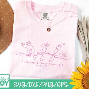 May include: A light pink t-shirt with a line drawing of three birds perched on a branch. The text "I will sing of the goodness of god PSALM 89:1" is printed below the birds. A gold necklace is draped on the shirt.