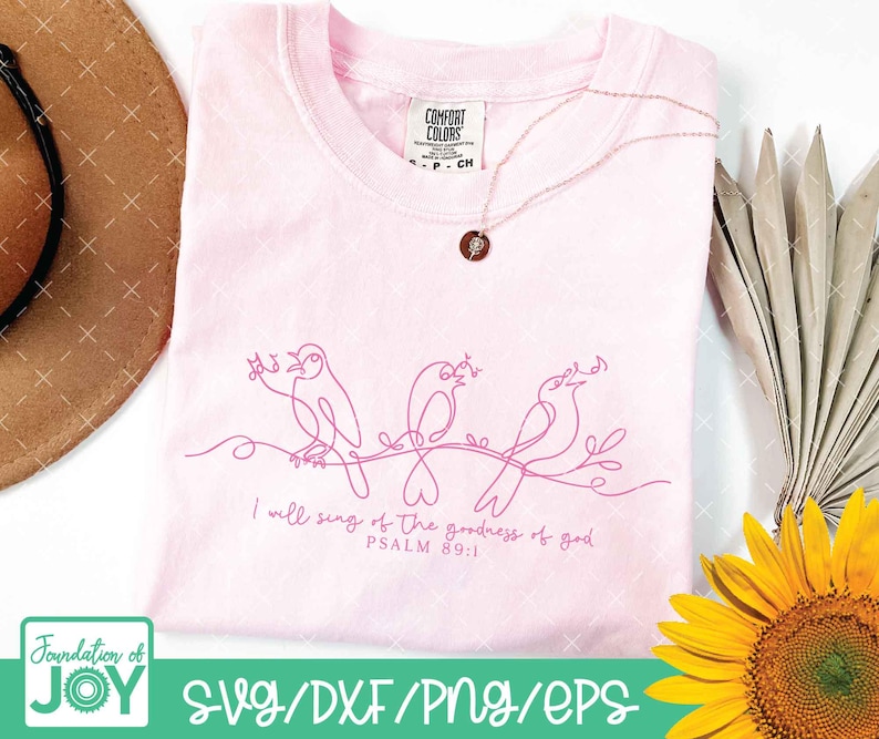 May include: A light pink t-shirt with a line drawing of three birds perched on a branch. The text "I will sing of the goodness of god PSALM 89:1" is printed below the birds. A gold necklace is draped on the shirt.