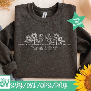 May include: Dark gray sweatshirt with a white line drawing of sunflowers, butterflies, and a sun. The text "When you can't find the sunshine, be the sunshine" is below the drawing. A green star with "WHITE PNG INCLUDED" is in the upper right.