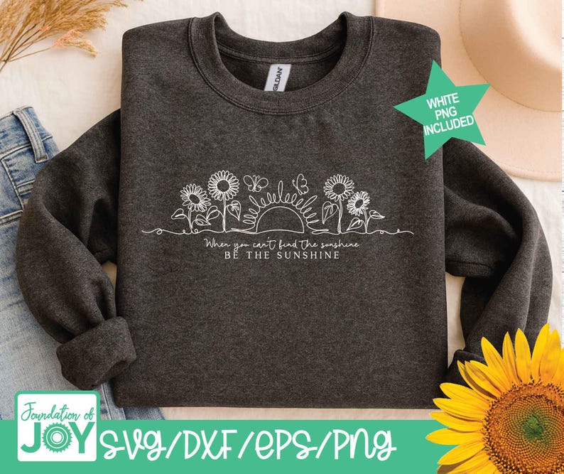 May include: Dark gray sweatshirt with a white line drawing of sunflowers, butterflies, and a sun. The text "When you can't find the sunshine, be the sunshine" is below the drawing. A green star with "WHITE PNG INCLUDED" is in the upper right.