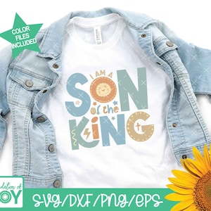 May include: A white t-shirt with the text "I am a Son of the King" in colorful letters with a lion graphic. The text is in a playful font with stars and a cross graphic. The t-shirt is displayed on a denim jacket and a sunflower.
