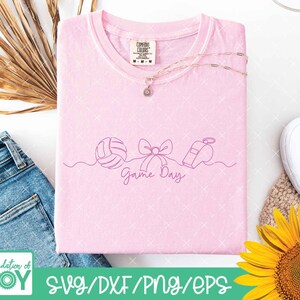 May include: A light pink t-shirt with a line drawing of a volleyball, bow, and whistle, with the words "Game Day" in cursive. The shirt is folded and has a Comfort Colors tag. A gold necklace with a pendant is on top of the shirt.