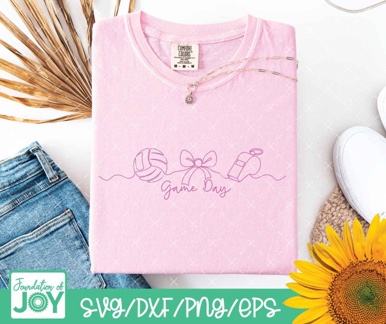 May include: A light pink t-shirt with a line drawing of a volleyball, bow, and whistle, with the words "Game Day" in cursive. The shirt is folded and has a Comfort Colors tag. A gold necklace with a pendant is on top of the shirt.
