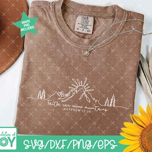 May include: A brown Comfort Colors t-shirt with a white mountain and sun graphic. The text "Faith can move mountains, Matthew 17:20" is printed below the graphic. A silver necklace and a sunflower are also visible.
