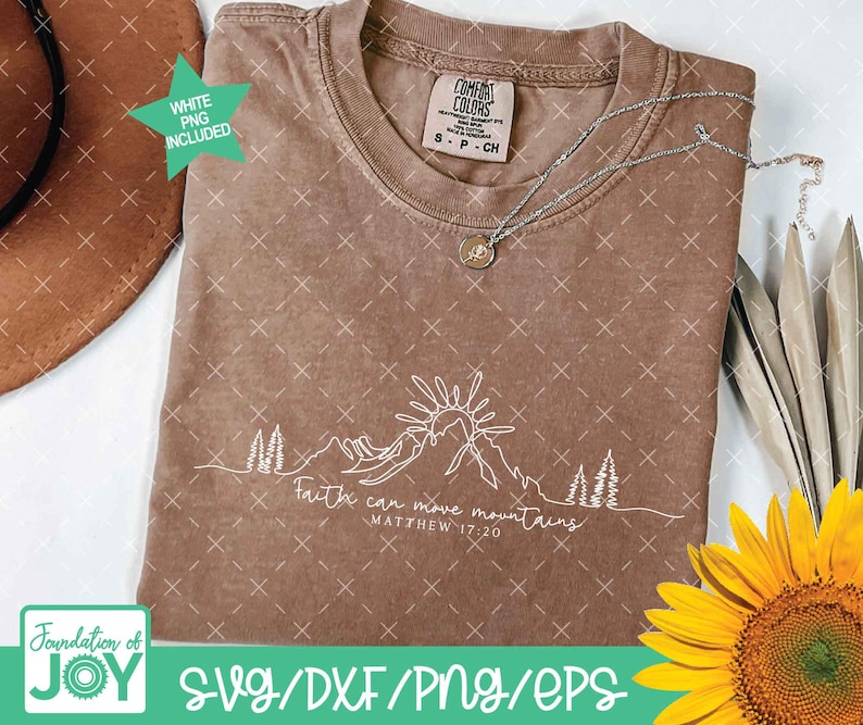 May include: A brown Comfort Colors t-shirt with a white mountain and sun graphic. The text "Faith can move mountains, Matthew 17:20" is printed below the graphic. A silver necklace and a sunflower are also visible.
